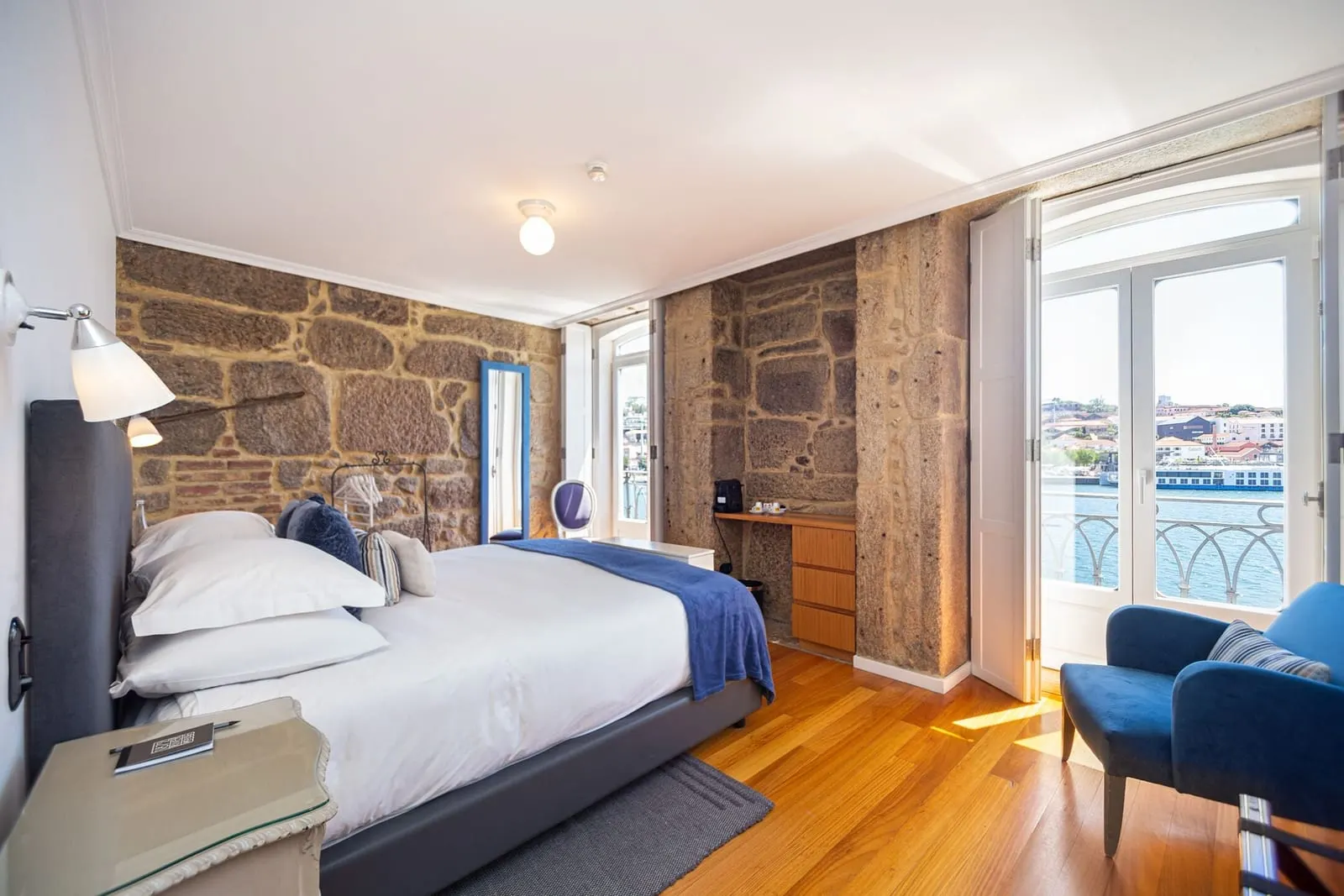 Superior Room with River View at 1872 River House: the bed, stone walls, a blue armchair and balcony doors open to the Douro