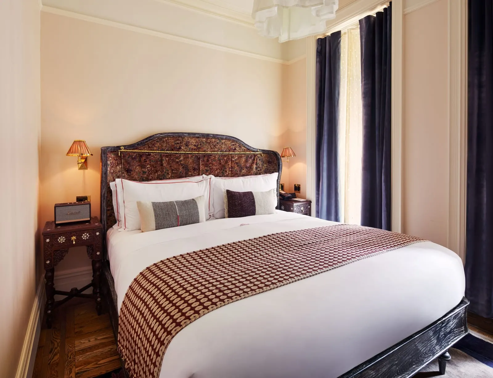 Petite Chelsea at Hotel Chelsea: a bed with a carved patterned headboard and a red and cream throw, between two small carved bedside tables, with a tall curtained window beside it