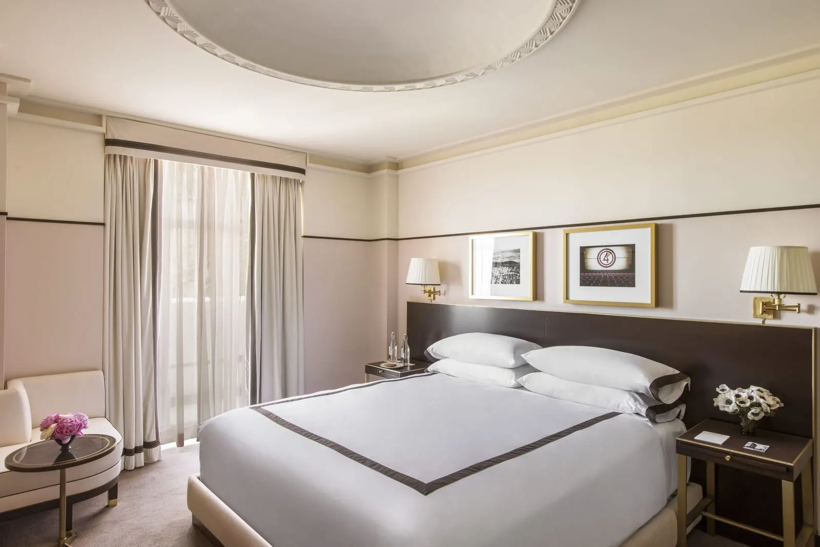 Deluxe King at Sunset Tower Hotel: a king bed with a dark headboard and brass wall lamps, a cream chaise with a round side table and a tall window with sheer curtains