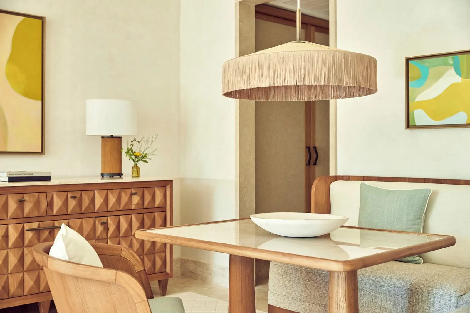 Sea-View Suite im Four Seasons Resort Mallorca at Formentor: ein Esstisch aus Holz mit einer gepolsterten Bank und einem Stuhl unter einer Hängeleuchte mit Fransen, neben einem geschnitzten Holzsideboard