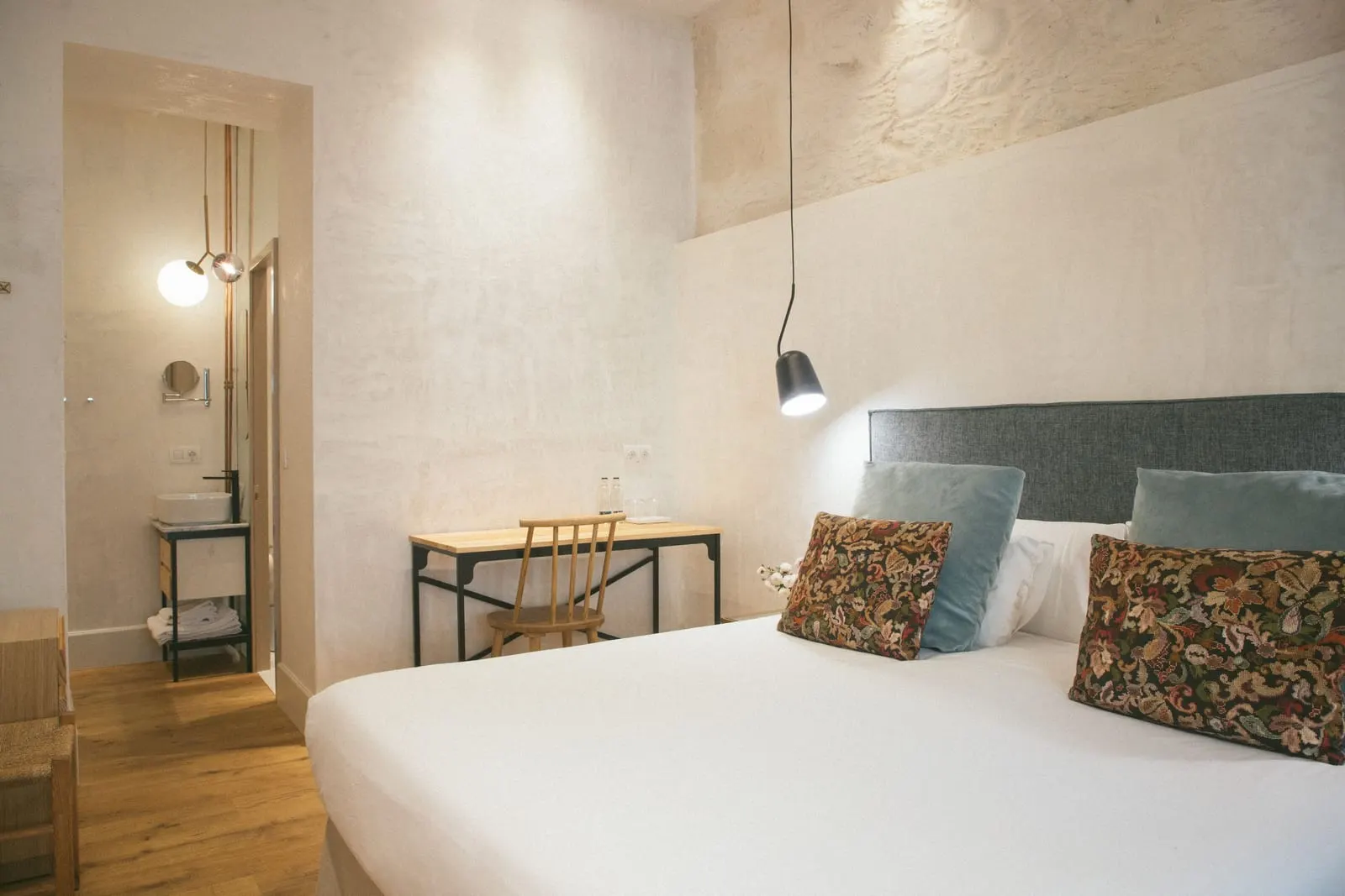 Deluxe Exterior at Hotel Veintiuno: a double bed with a grey headboard and floral cushions under a black pendant lamp, a wooden desk and chair, and a washbasin alcove beyond