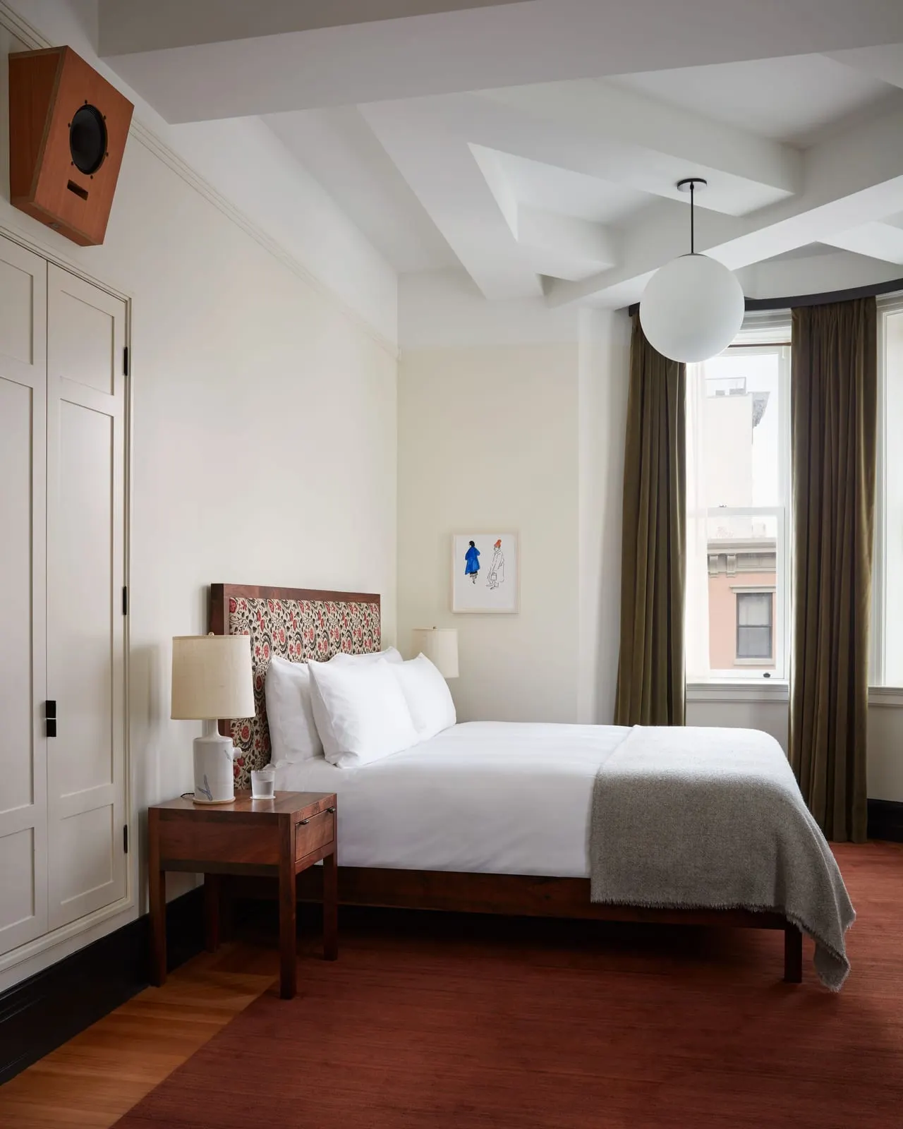 Corner King at Nine Orchard: a bed with a patterned fabric headboard and a grey wool throw beside the curved window bay, with a panelled cupboard on the left