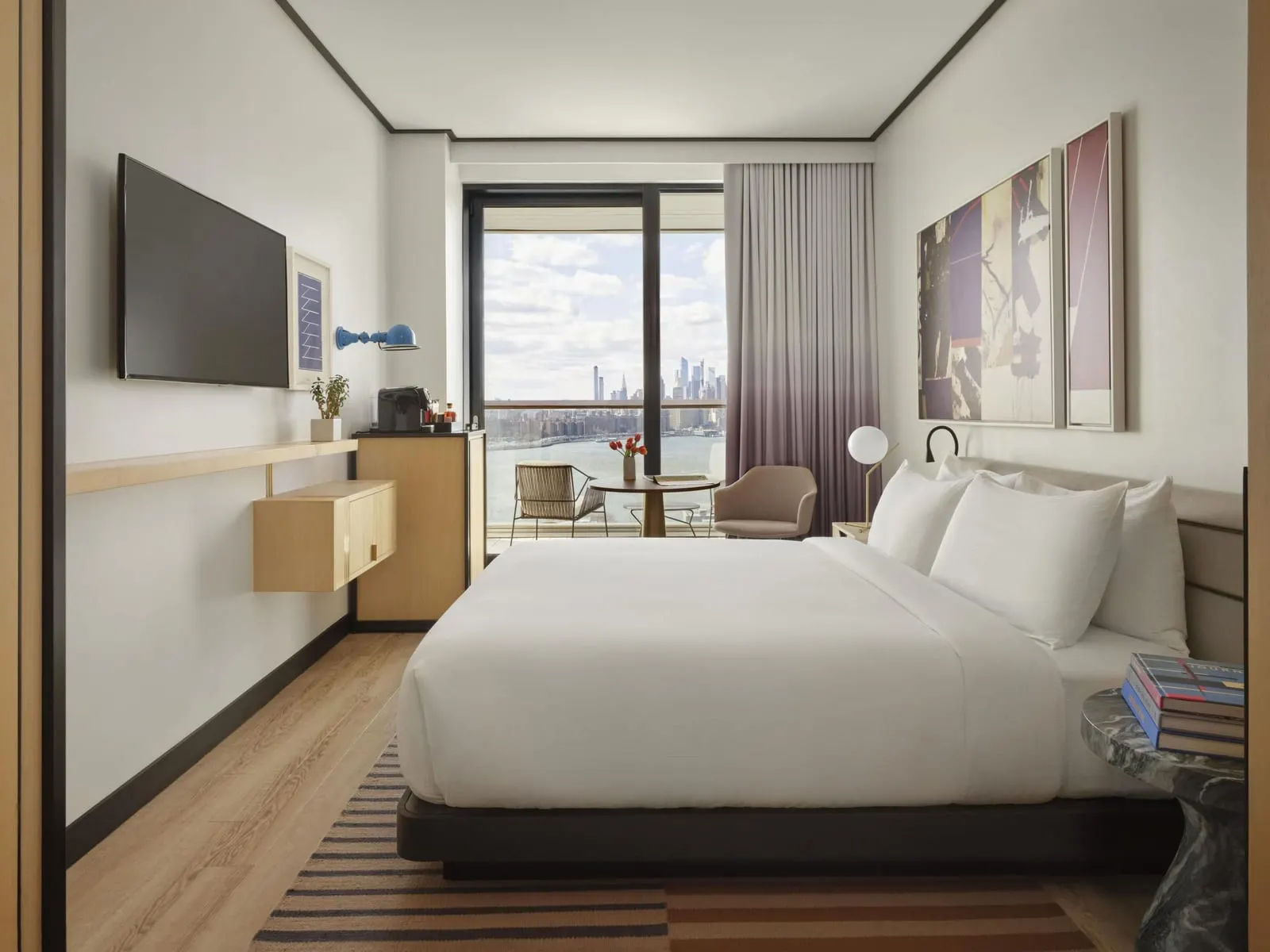 Manhattan View King with Balcony at The William Vale: a bed with white linen below two framed abstract paintings, a wall-mounted television on the opposite wall, and an armchair by the window with the river and the Manhattan skyline beyond