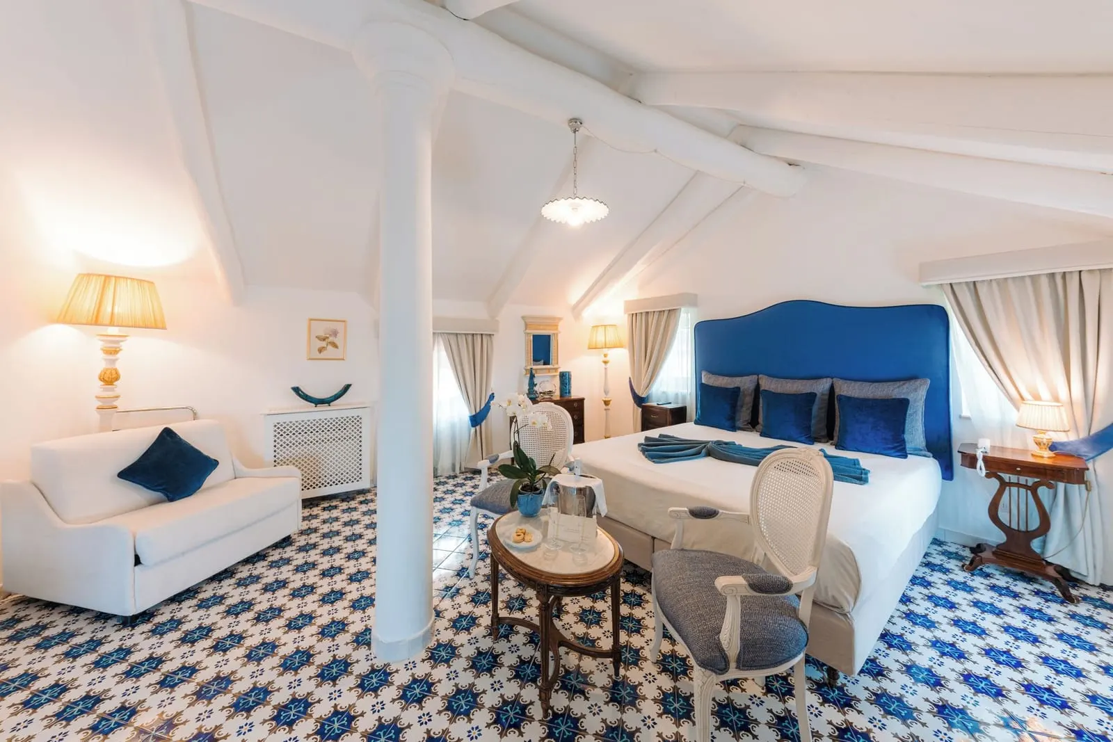 Junior Suite ORTENSIA at Palazzo Pascal: a double bed with a blue velvet headboard, a white sofa and a sitting table under a beamed ceiling