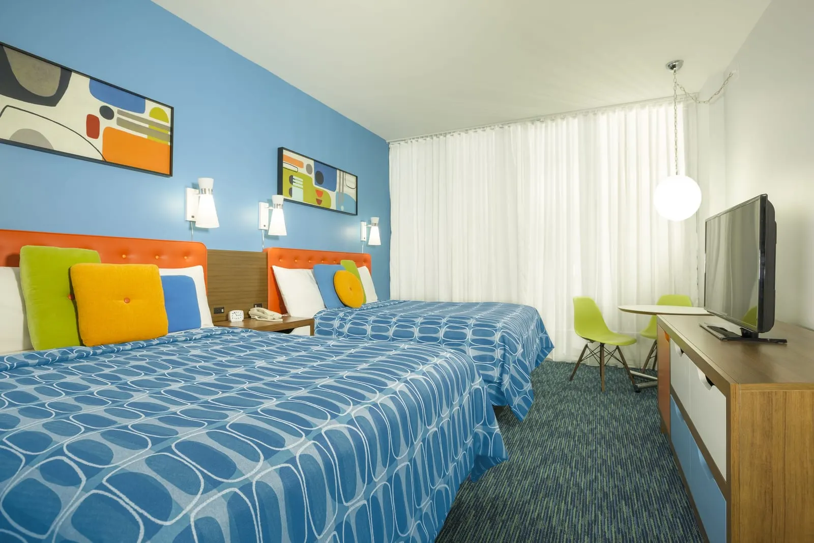 Standard 2 Queen Beds at Universal Cabana Bay Beach Resort: two queen beds with orange headboards against a blue wall, a dresser with a television and a small round table with two green chairs by the curtained window