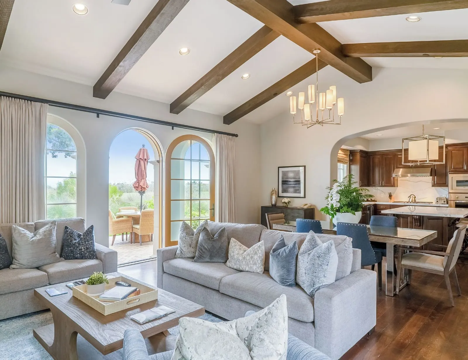 Three-Bedroom Ocean Villa at Terranea Resort: an open living and dining room with grey sofas, a kitchen beyond and arched windows under a beamed ceiling