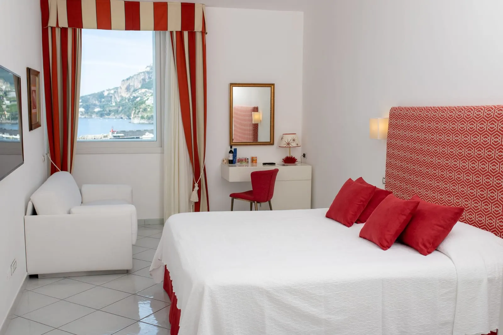 Tripla Standard（Hotel Marina Riviera）：赤いクッションのダブルベッド、白いアームチェア、ドレッシングテーブルがあり、縞のカーテンの窓は湾に面している