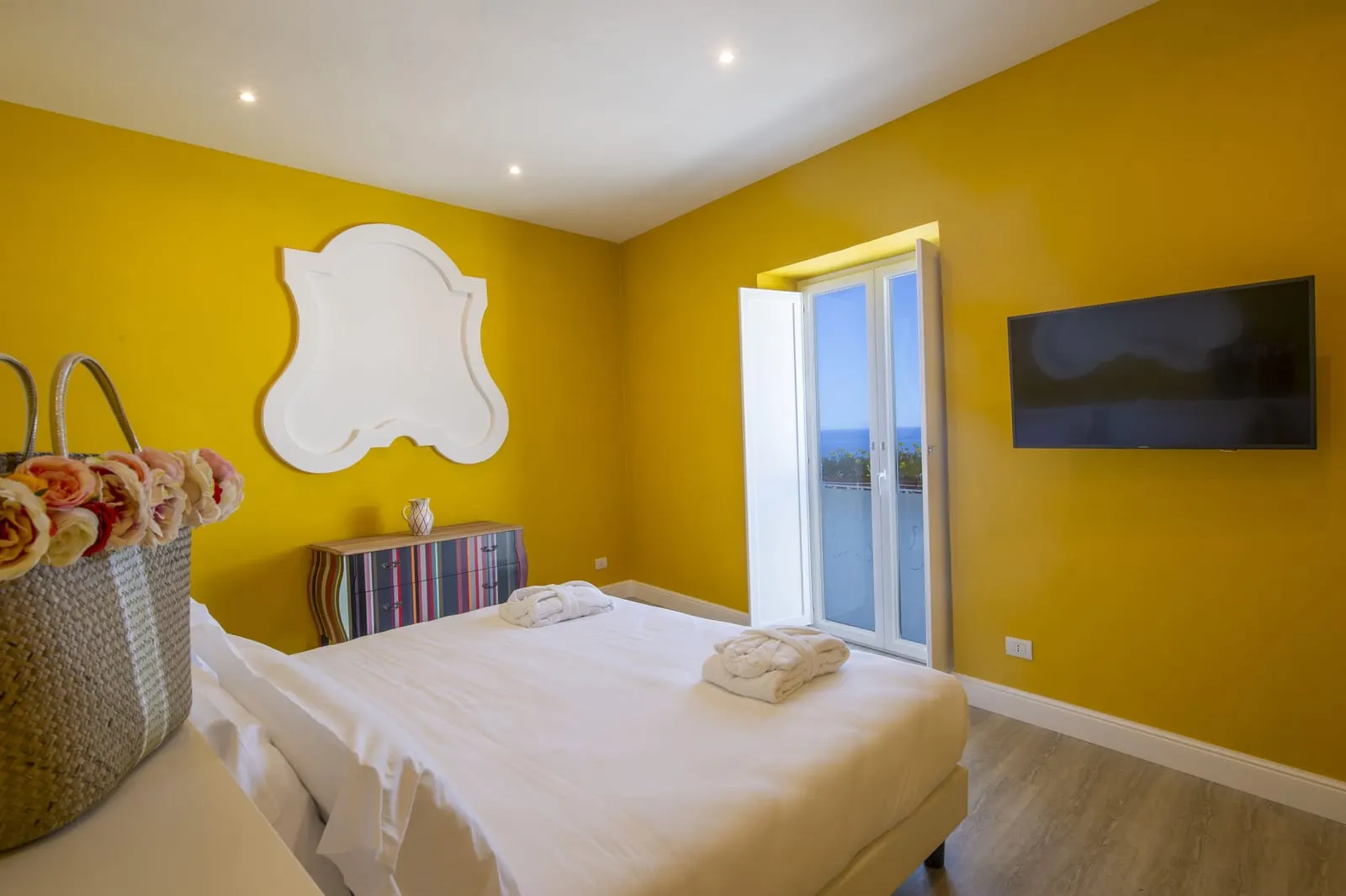 Apartment Suite Melina all'Hotel Tramonto d'Oro: un letto matrimoniale in una camera dalle pareti gialle con una cassettiera a righe, un televisore a parete e una porta di vetro.