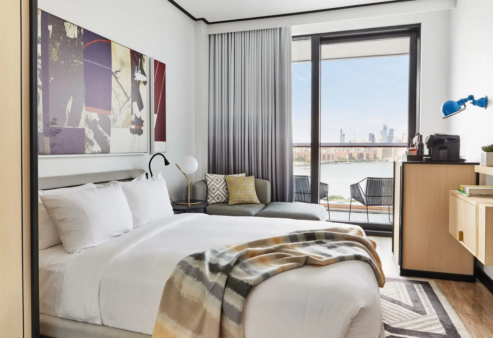 Manhattan View Queen with Balcony at The William Vale: a bed with a striped throw under a large purple and cream collage painting, a grey sofa by the window, an oak cabinet with a coffee machine below a blue wall lamp, and a glass balcony door with two chairs outside and the river and the Manhattan skyline beyond
