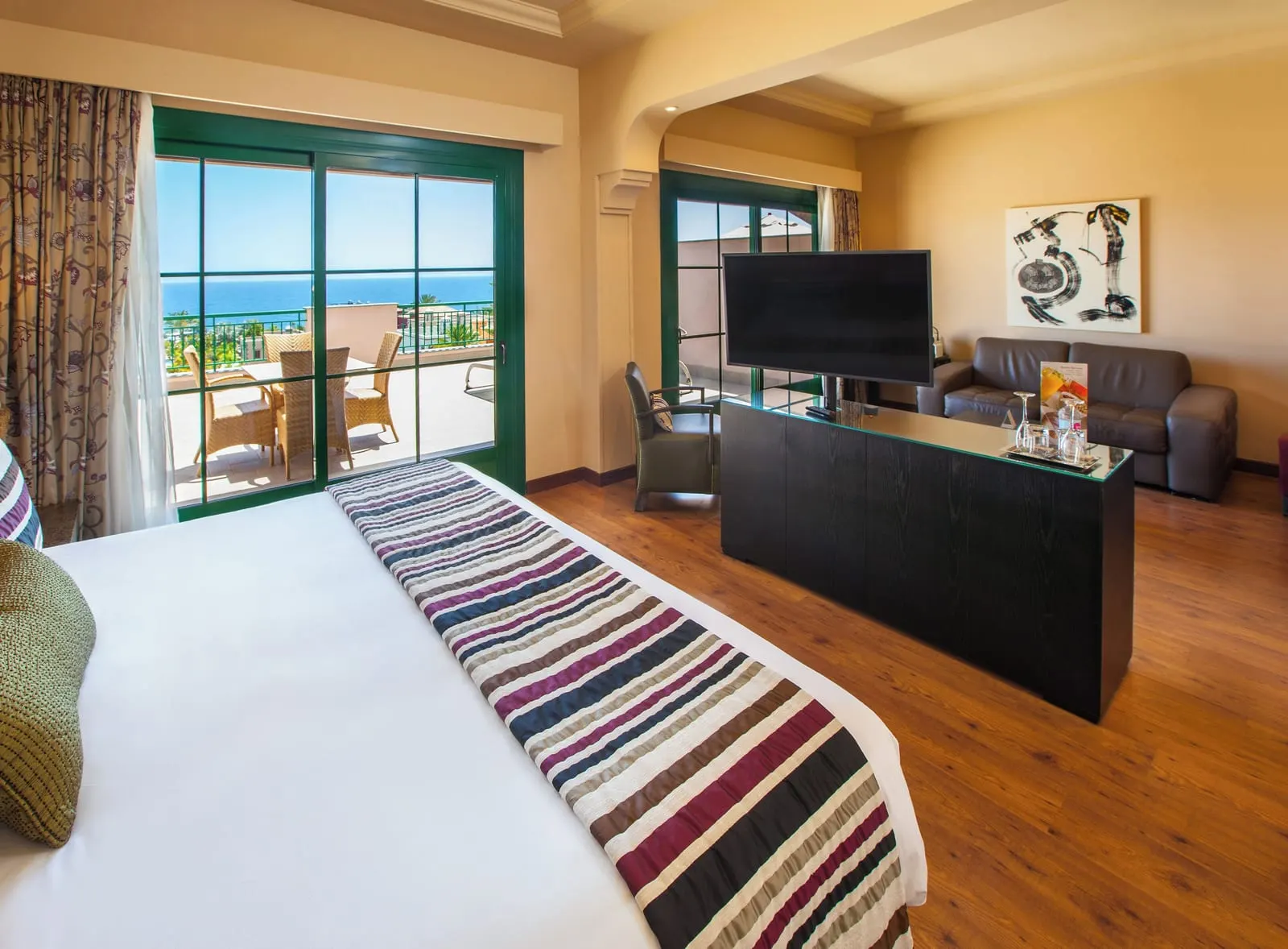 Double Deluxe View at Lopesan Villa del Conde Resort & Thalasso: a king bed with a striped runner facing a television cabinet, a leather sofa and green-framed glass doors to a terrace with a table and wicker chairs