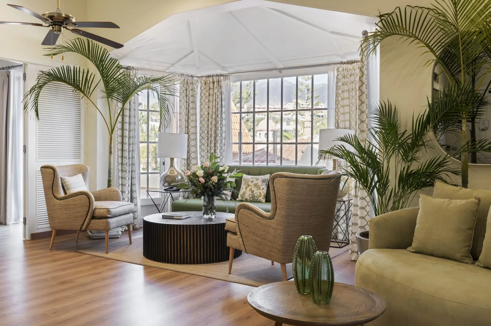 Suite at Bahía del Duque: the living room, with armchairs, a sofa and palms in front of a glazed bay window