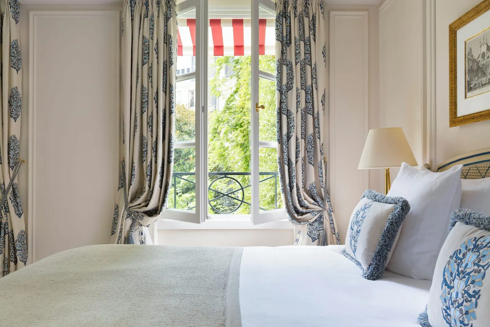 Junior Suite with garden view at Le Bristol Paris: the bed beside an open window with green trees outside, framed by blue patterned curtains under a striped awning
