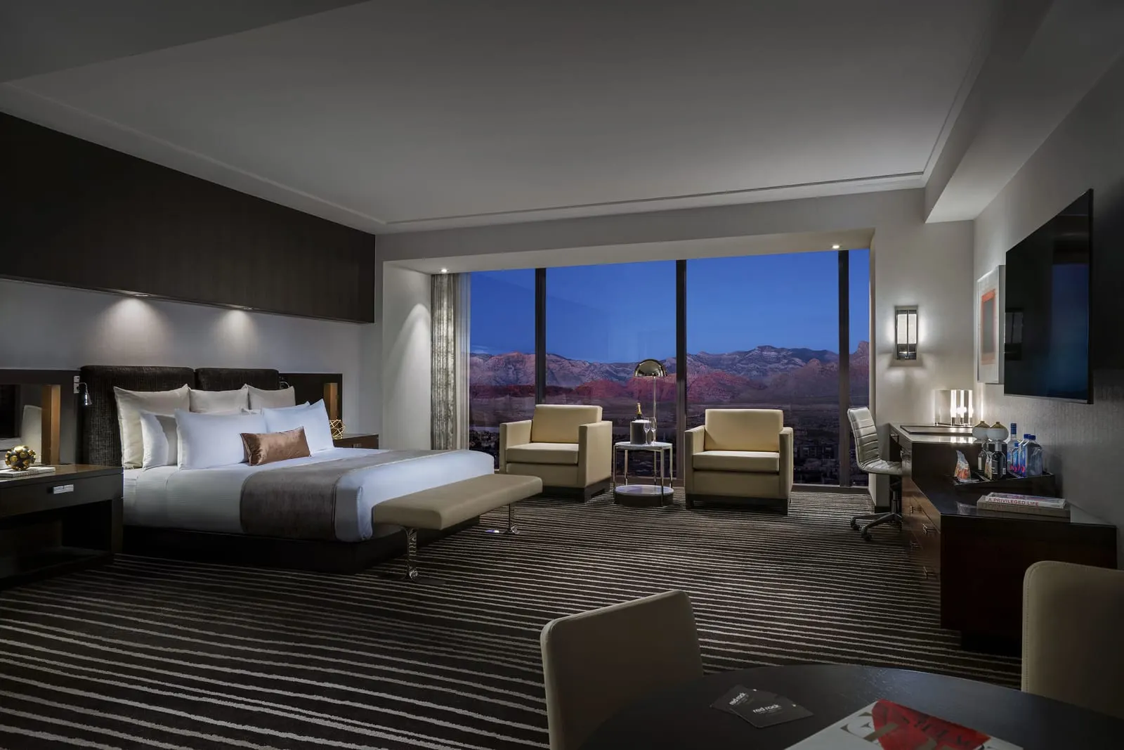 Executive King Corner Room at Red Rock Casino Resort & Spa: a king bed with an upholstered bench at its foot, two armchairs by a floor-to-ceiling window, a desk and dresser along the right-hand wall, and a round table with chairs in the foreground