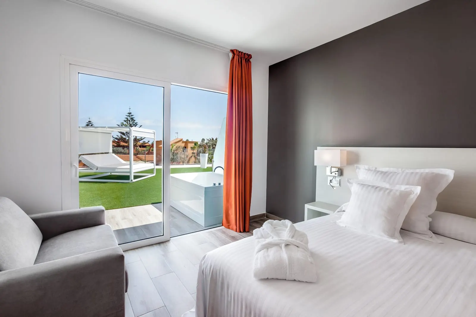 Deluxe with Hot Tub at Barceló Corralejo Sands: a bed with bathrobes and glass doors onto a grass terrace with its own outdoor tub and a daybed.
