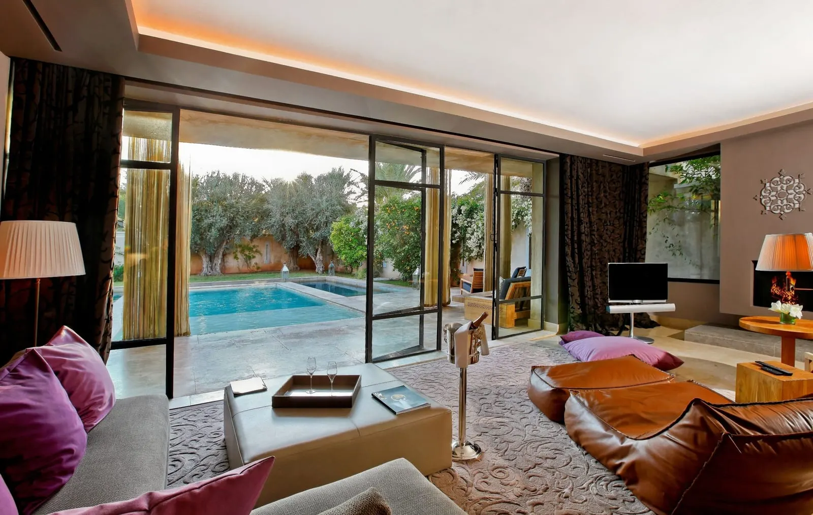 Pool Villa at Palais Namaskar: a living room with low sofas and glass doors open onto a private pool among olive trees