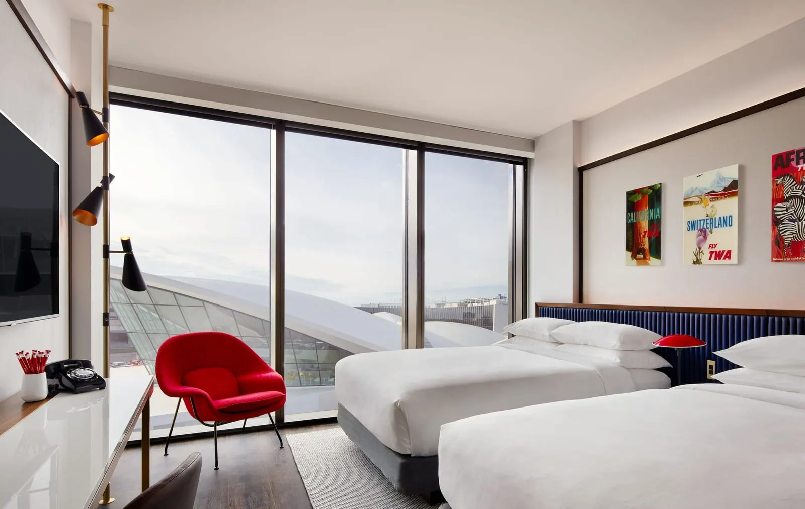 Deluxe Double Queen With Historic TWA View at TWA Hotel: two queen beds and a red armchair beside floor-to-ceiling windows that frame the curved white roof and glass wall of the TWA Flight Center