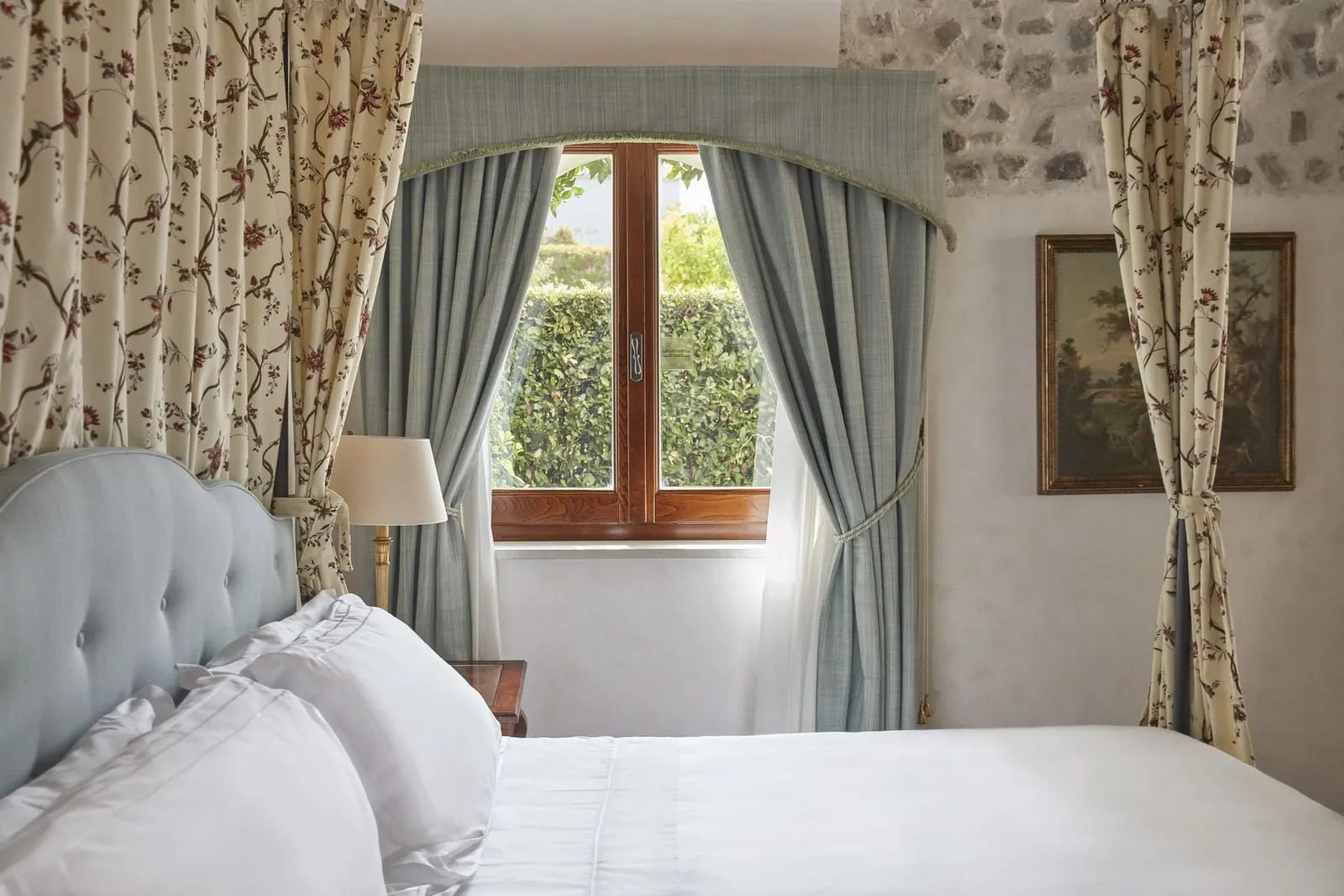 Superior Room with Garden at Caruso, A Belmond Hotel: a bed with a pale blue headboard beside a window onto a green hedge, with floral curtains and a framed landscape painting