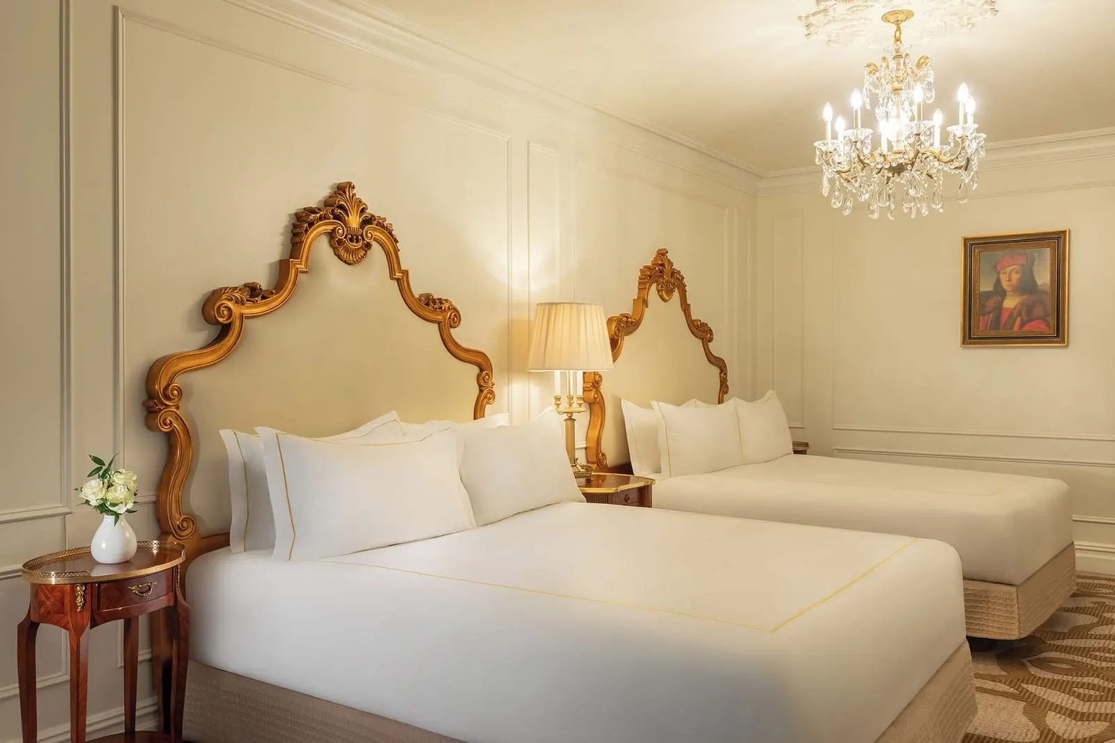 Deluxe Two Queens at The Plaza: two queen beds with gilded headboards, a lamp between them and a small round table with a vase of white roses