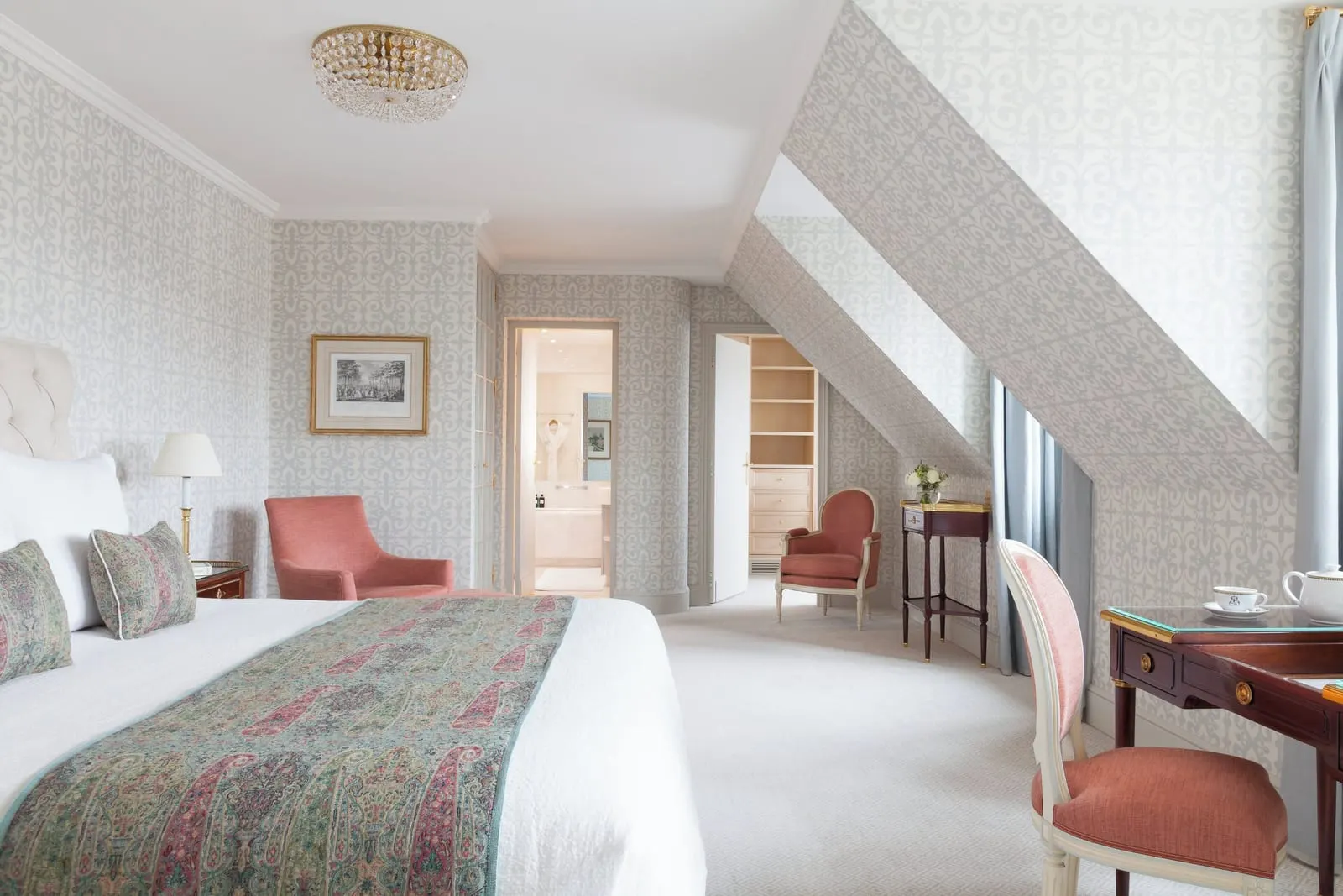 Suite 1925 at Le Bristol Paris: a bed with a paisley throw under a sloping ceiling, coral armchairs and a writing desk, with a bathroom and shelving through two doorways