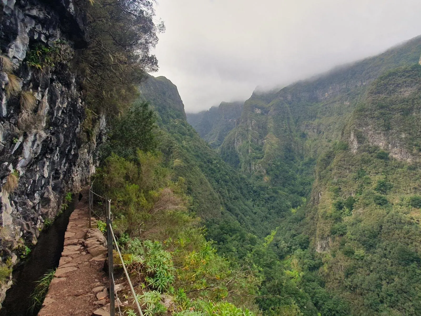 A stone path beside the levada channel, cut into a cliff above a deep green valley under low cloud
