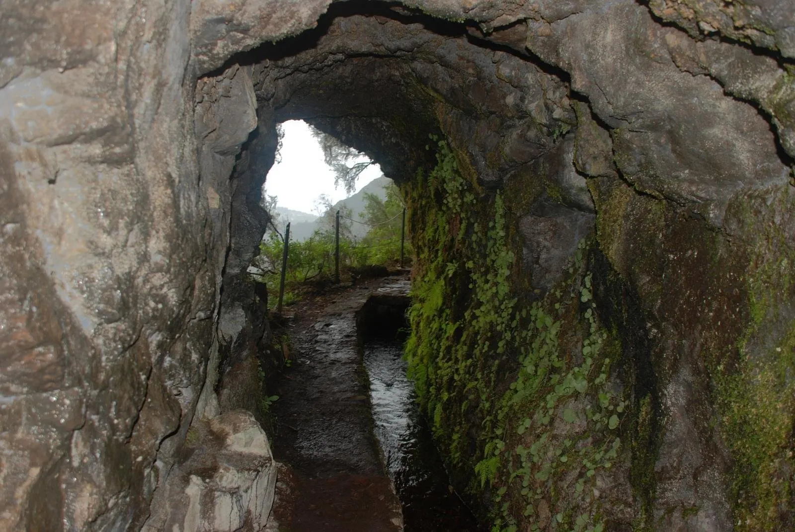 Inside a rock tunnel on the levada, with water running in the channel and daylight at the far end
