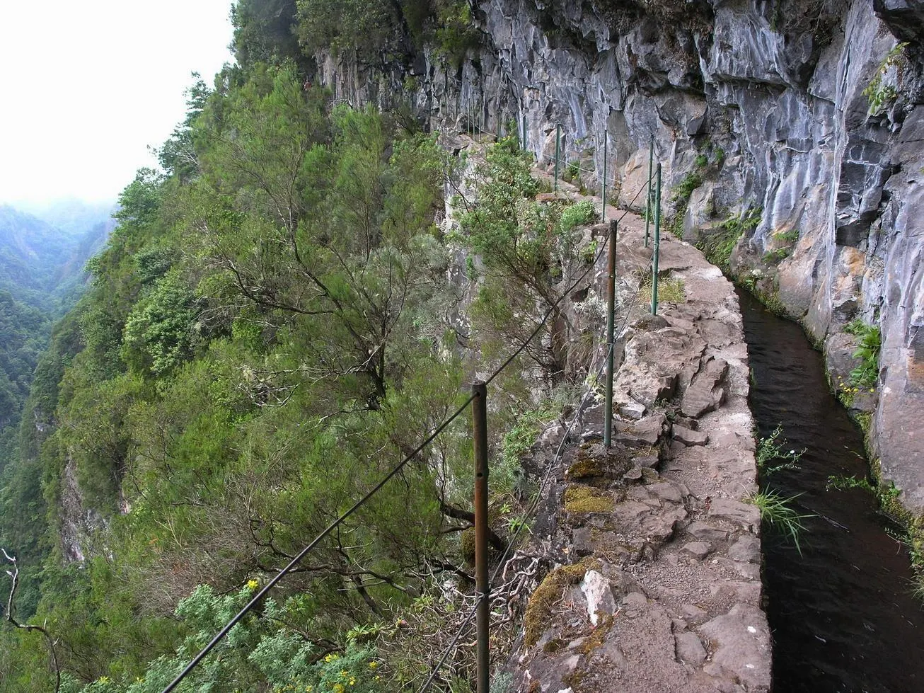 The levada walkway along the foot of a grey rock face, with a wire fence on the valley side