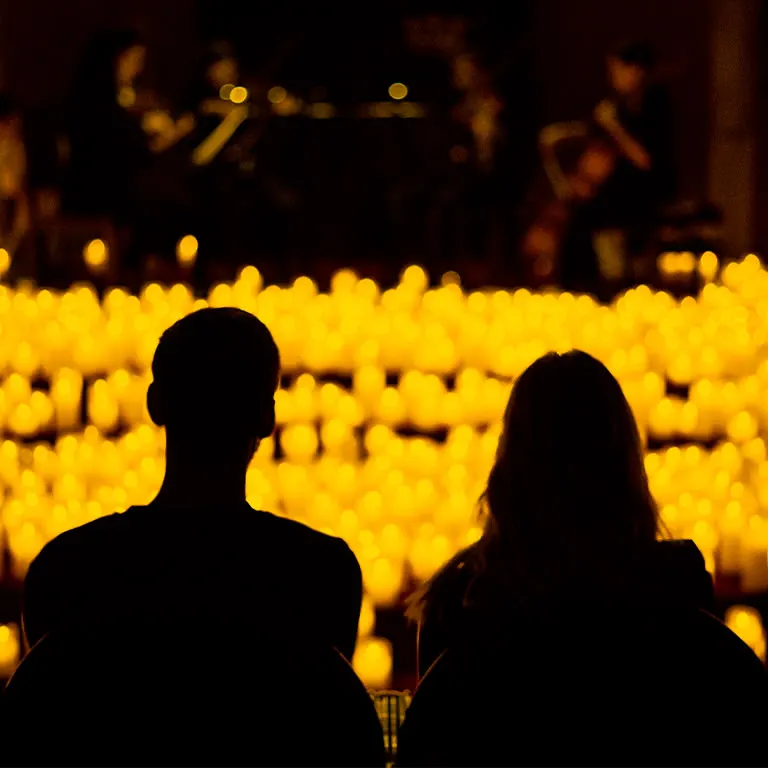 Two concertgoers seen from behind, facing a string quartet on a stage beyond rows of flameless candles at a Candlelight concert