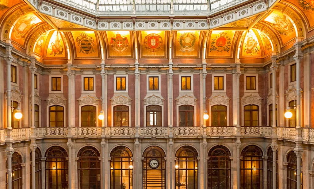 The Pátio das Nações inside the Palácio da Bolsa: two storeys of arched galleries under a glass roof, with painted coats of arms around its base, lit warm in the evening