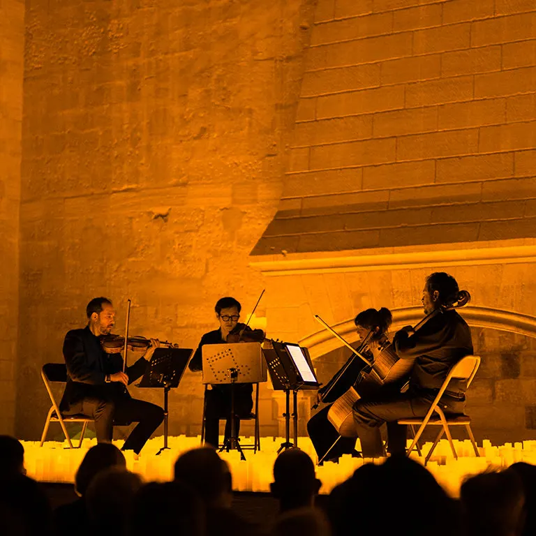 Four string players perform behind a bank of candles against a tall stone wall, the heads of the audience in the foreground