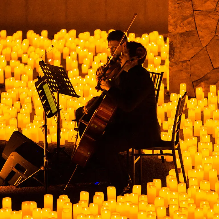 Candlelightコンサートで、石壁のそばで数百本のキャンドルに囲まれ、二人のチェリストが椅子に座って演奏している