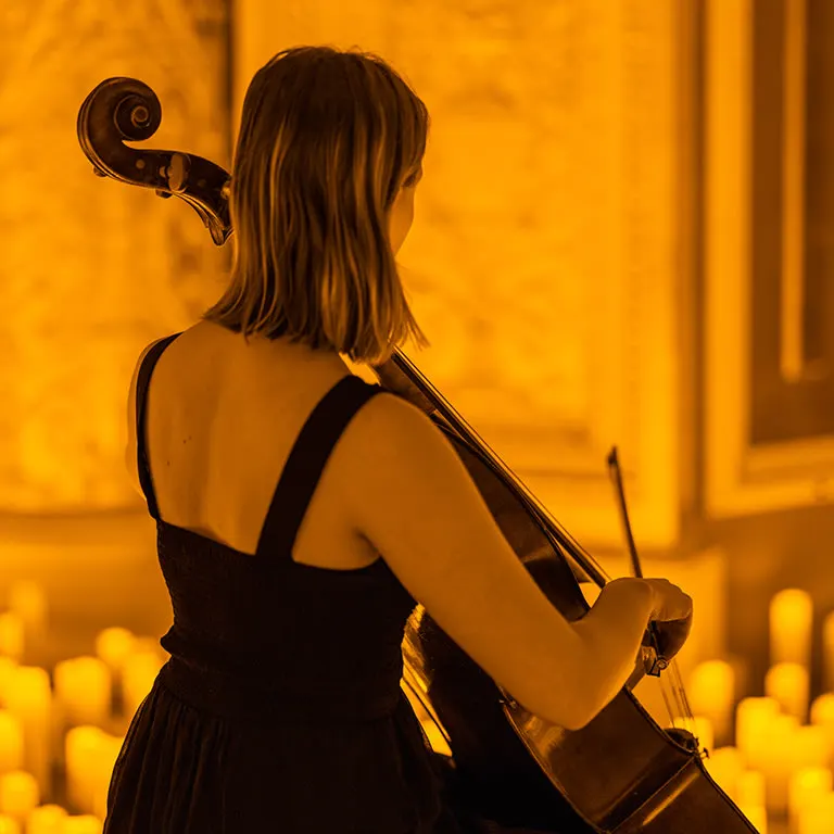 A cellist in a black dress plays with her back to the camera, candles glowing on the floor around her