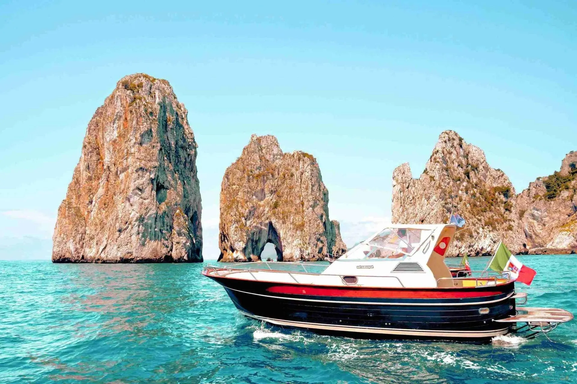 A small motorboat with an Italian flag passing the Faraglioni rocks off Capri, one of them pierced by a sea arch