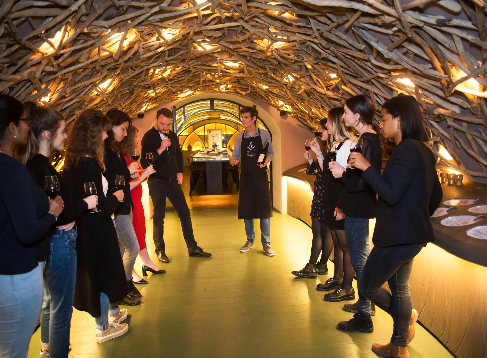 A guide in an apron holds up a bottle for a group of guests with glasses of red wine in a cellar tunnel roofed with lit vine branches