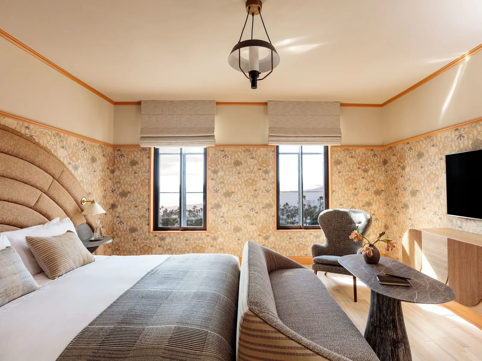 Landmark Grand King at Santa Monica Proper Hotel: a bed with an arched headboard, a curved bench, an armchair and a wall-mounted television beneath two windows