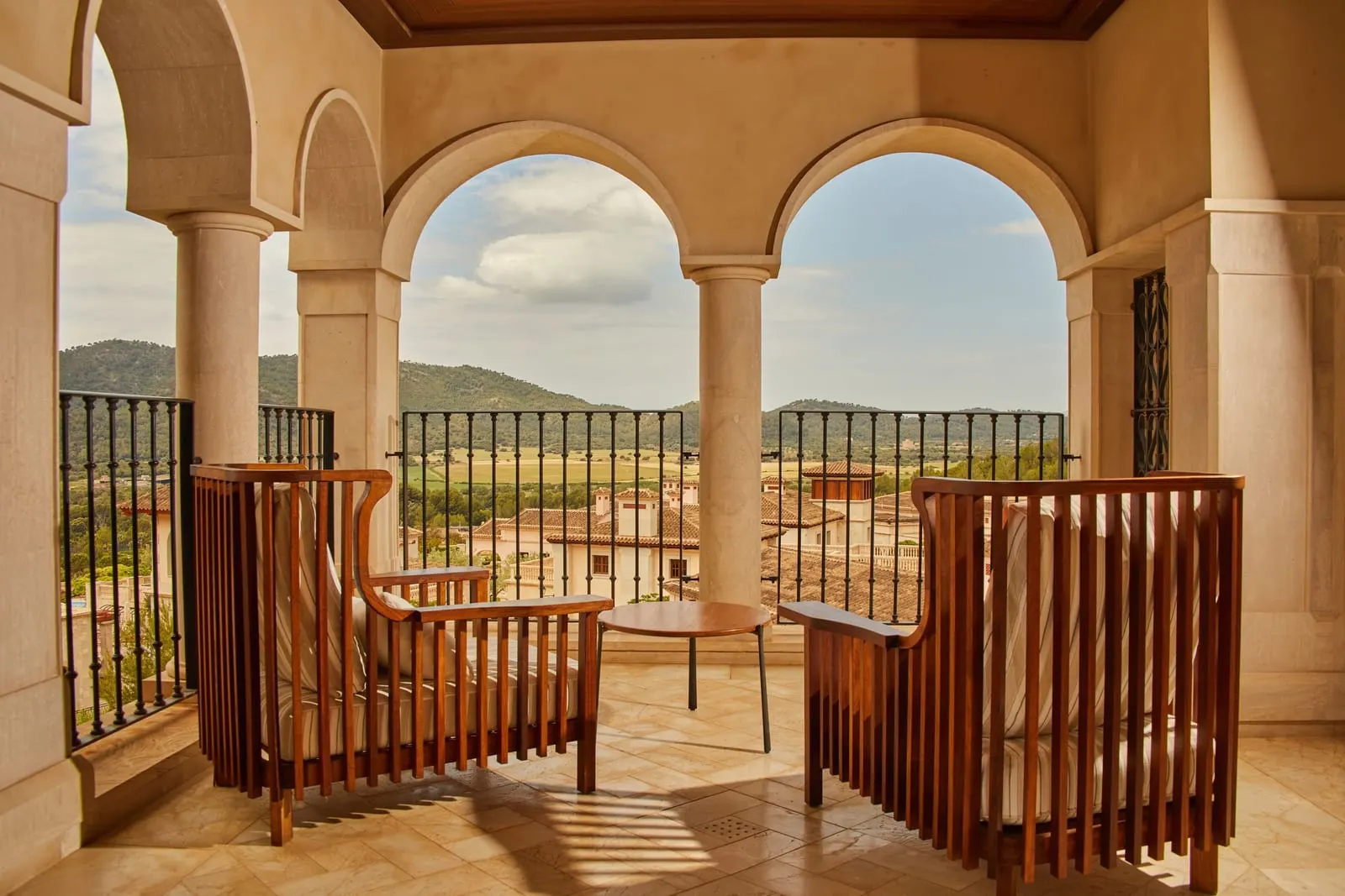 Junior Suite Valley View at Cap Vermell Grand Hotel: two timber armchairs on a stone terrace under arches, looking over the resort's rooftops to green hills