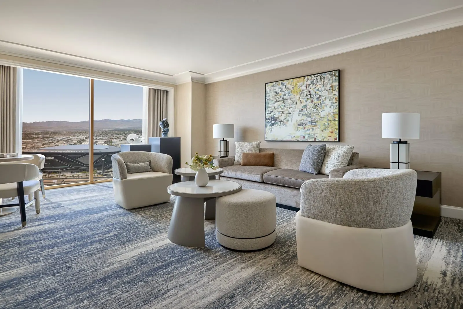 Stadium View One-Bedroom Suite at Four Seasons Hotel Las Vegas: a living room with a sofa, two armchairs and round tables under an abstract painting, and a floor-to-ceiling window onto Allegiant Stadium and the valley