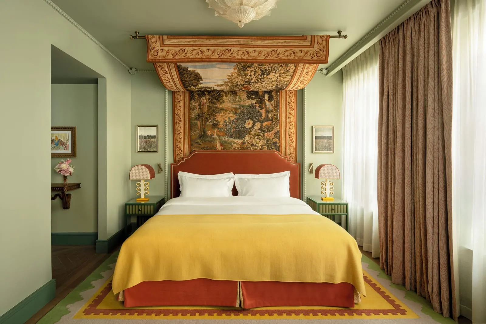 Prestige Room at Le Grand Mazarin: a bed with a rust velvet headboard and a yellow throw under a tapestry canopy, between matching bedside tables and lamps, with tall curtained windows to the right