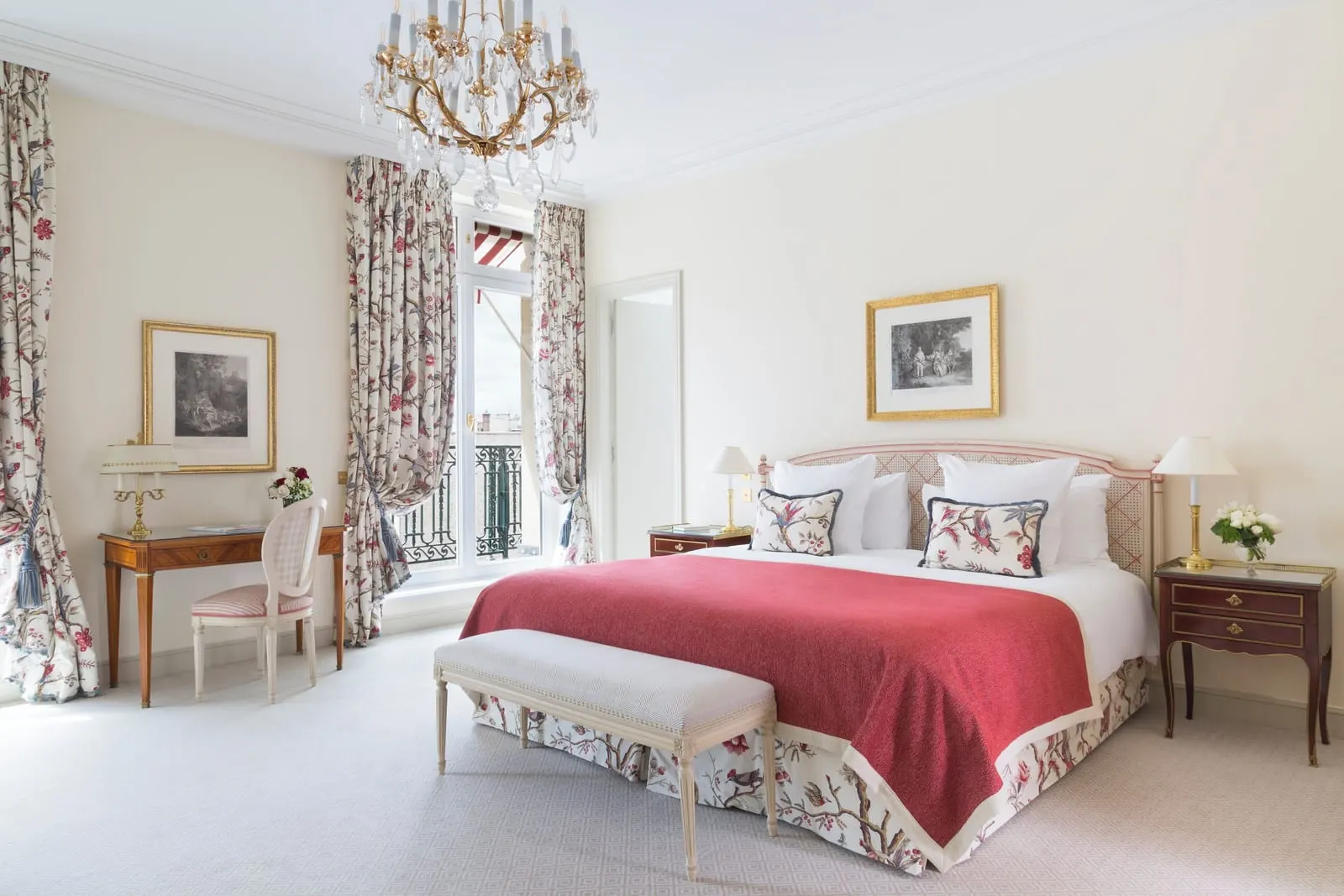 Suite Paris at Le Bristol Paris: a bed with a red throw and bird-patterned cushions, a writing desk and French windows with a wrought-iron balcony rail