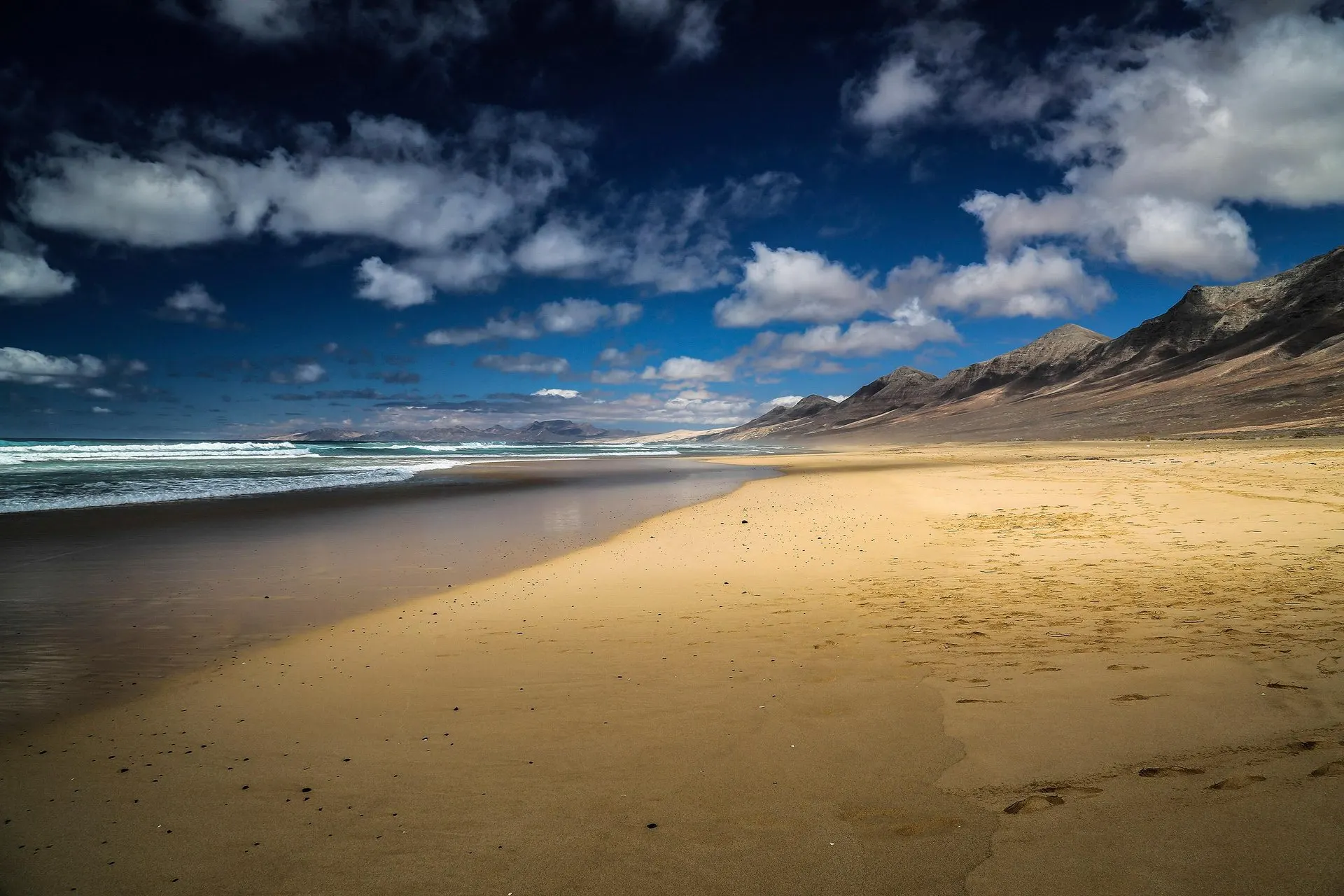 The wide, empty sand of Cofete beach under a dark blue sky with white clouds