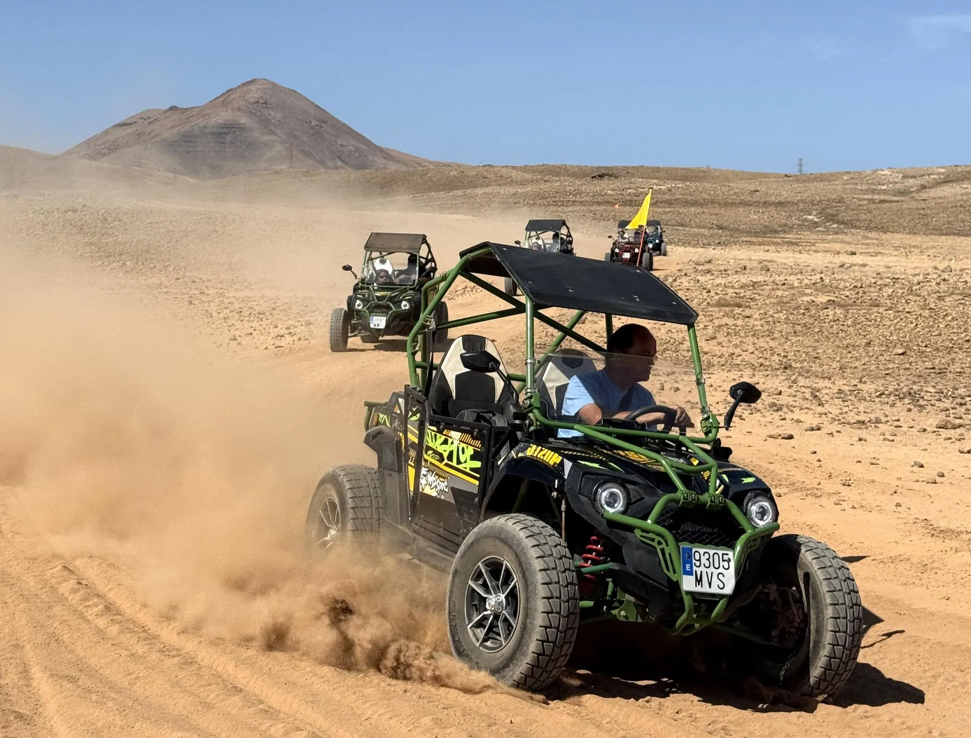 A green and black two-seat buggy kicking up dust on a dirt track, with more buggies behind and a volcanic cone on the skyline