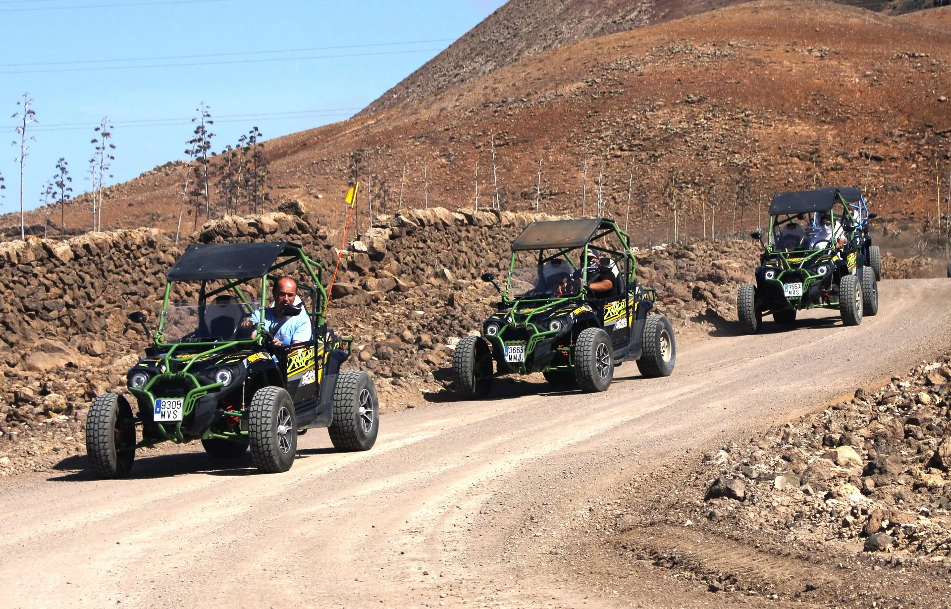 Three buggies in convoy on a gravel track beside a dry-stone wall, below a bare brown volcanic slope