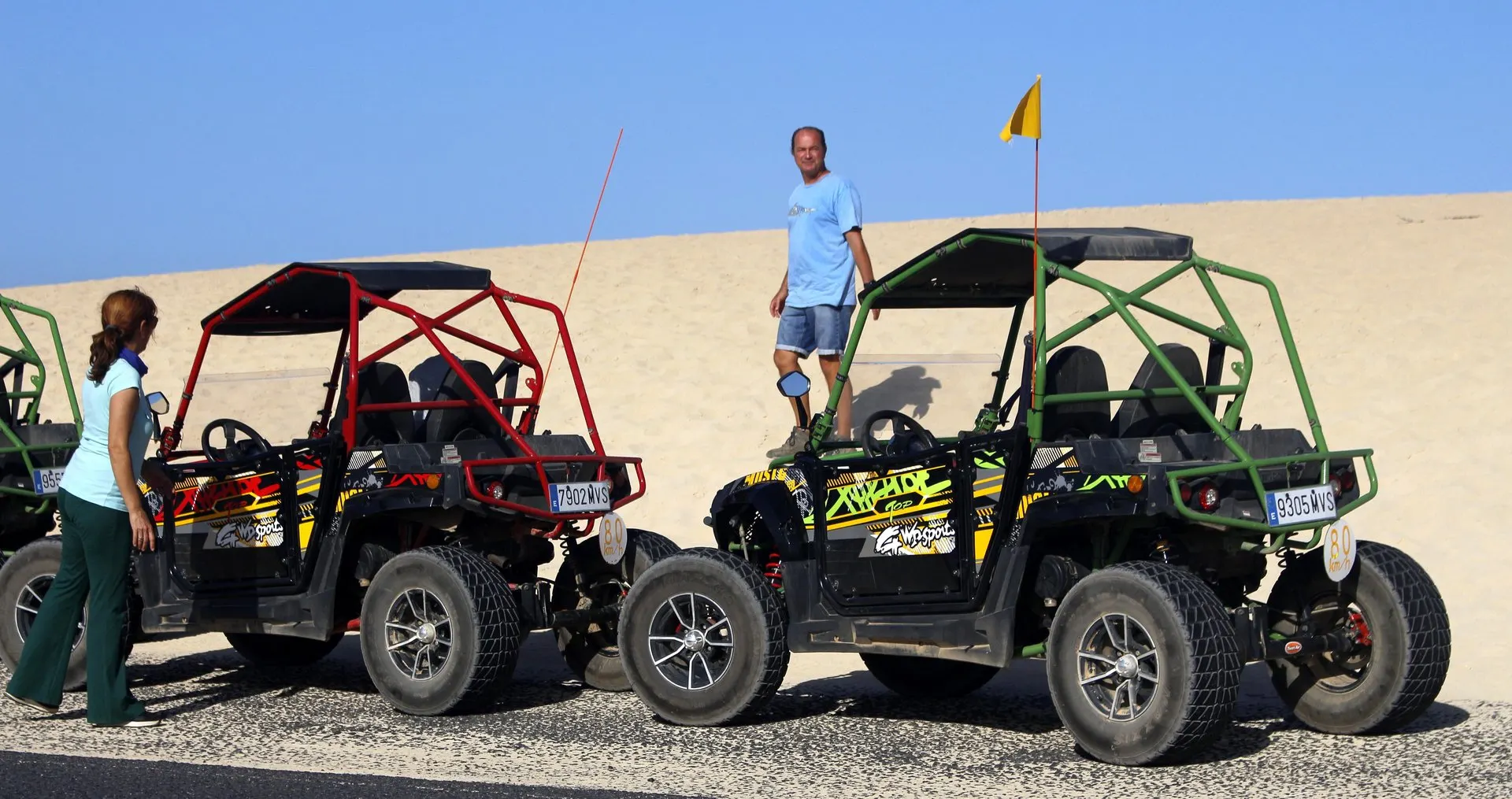 Buggies parked on the roadside at the foot of a white sand dune, with two people standing beside them
