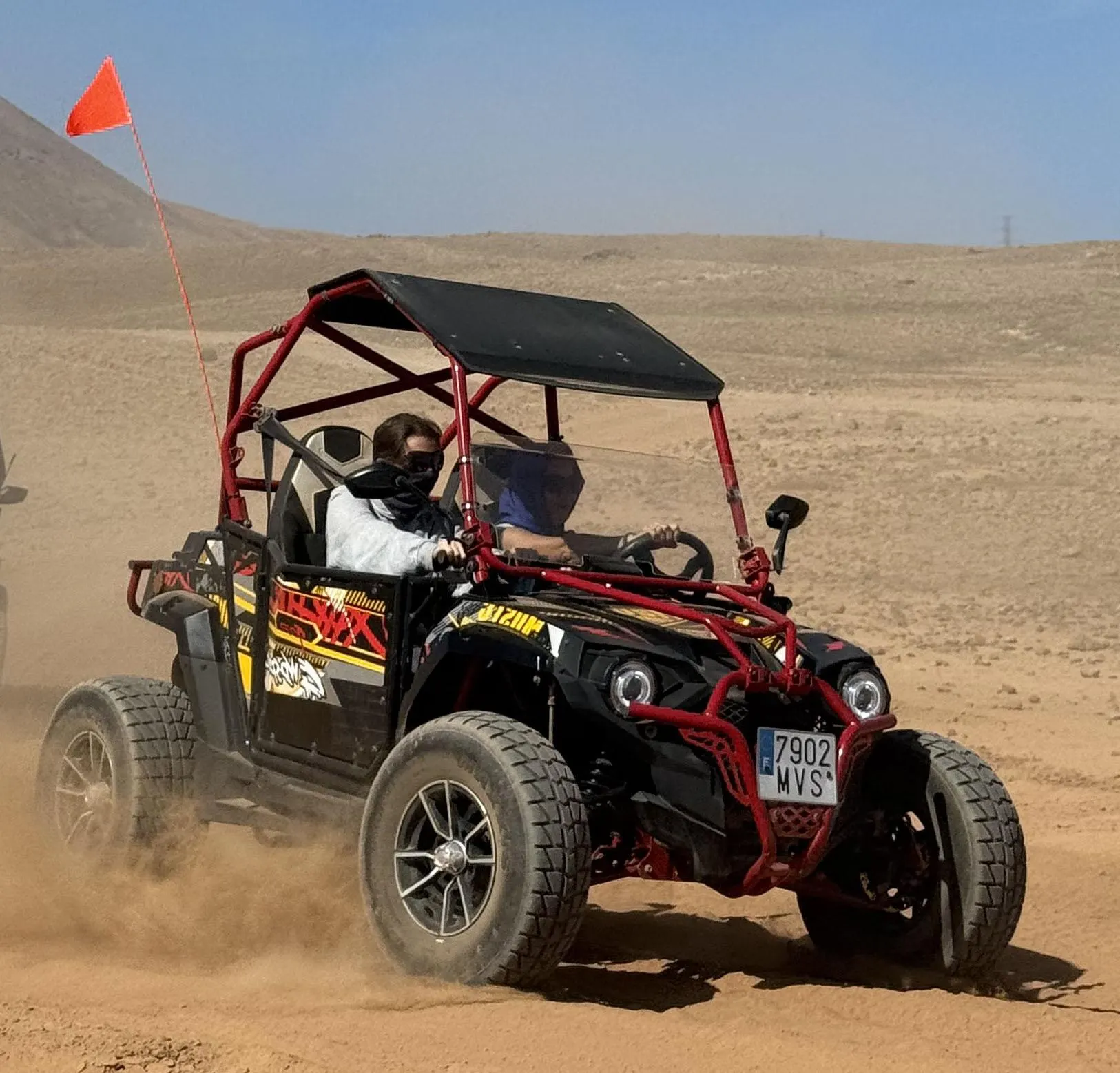 A driver and passenger with scarves over their faces in a red and black buggy on a dusty plain