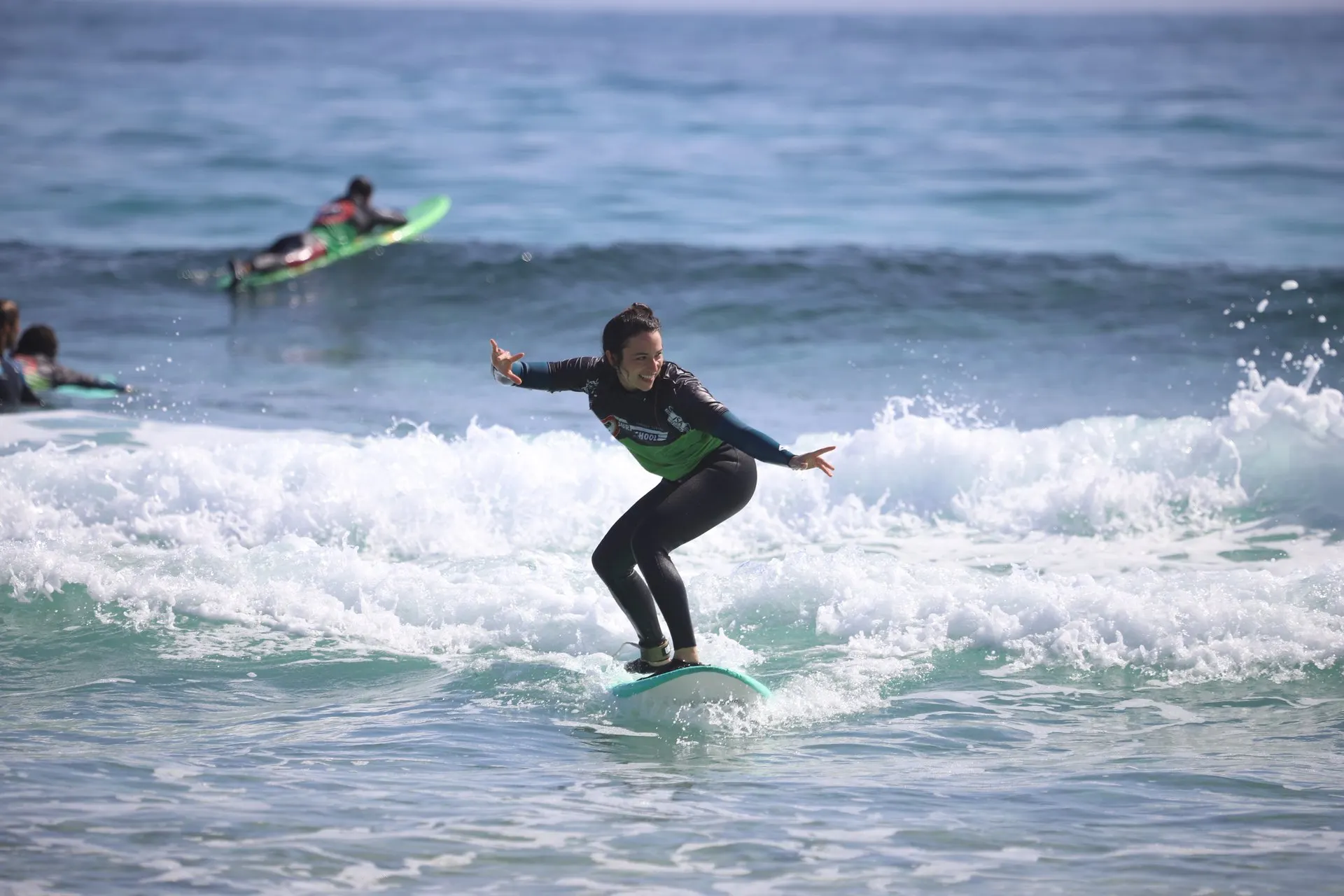 A learner in a wetsuit stands up on a soft surfboard in the whitewater, arms out, with other learners paddling behind