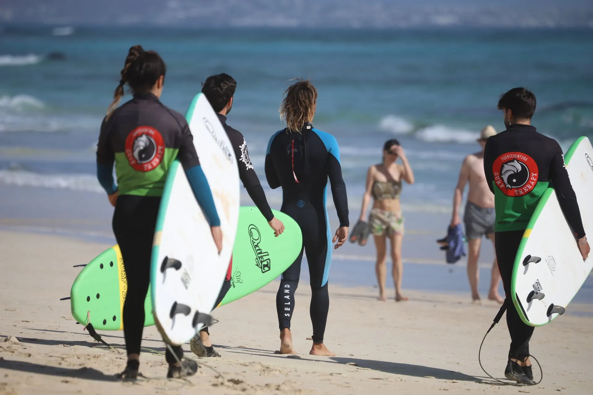 Four surfers in school rash vests and wetsuits carry soft boards down a sandy beach towards the waves