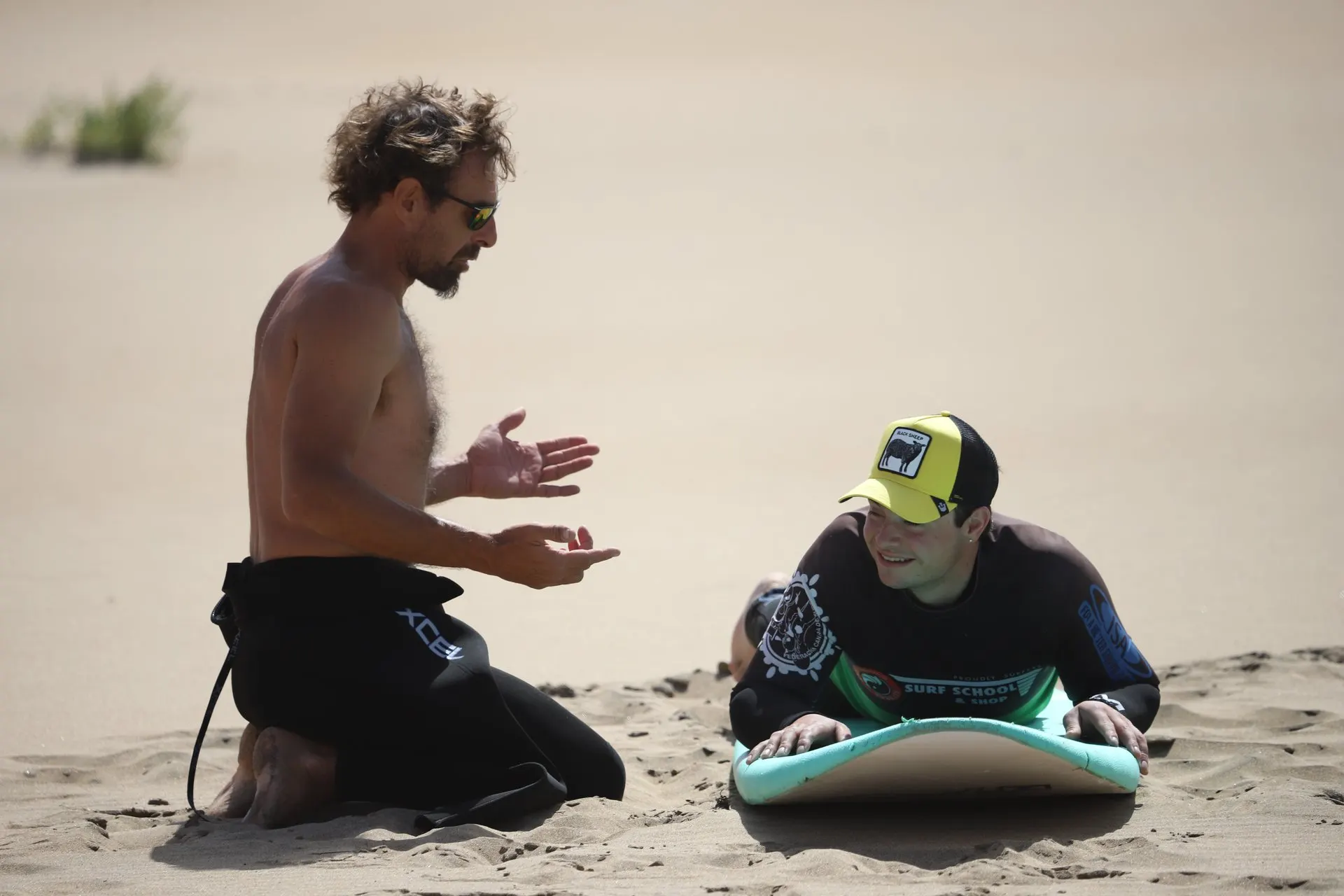 An instructor kneels on the sand explaining the stand-up to a learner lying on a surfboard