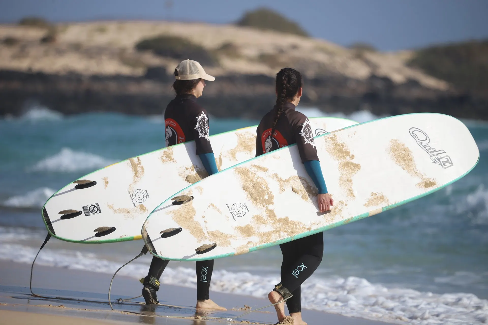 Two learners carry long soft boards along the shoreline, with low dunes and a rocky point behind