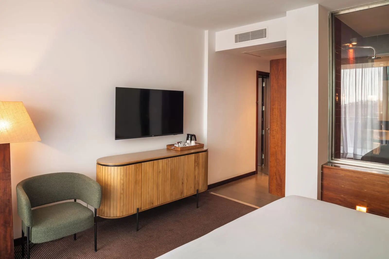 Double Deluxe with daily access to the Be Aloe Wellness at Salobre Hotel Resort & Serenity: a bedroom with a wall-mounted television above a wooden sideboard with a kettle tray, a green armchair and a glass panel into the rain shower