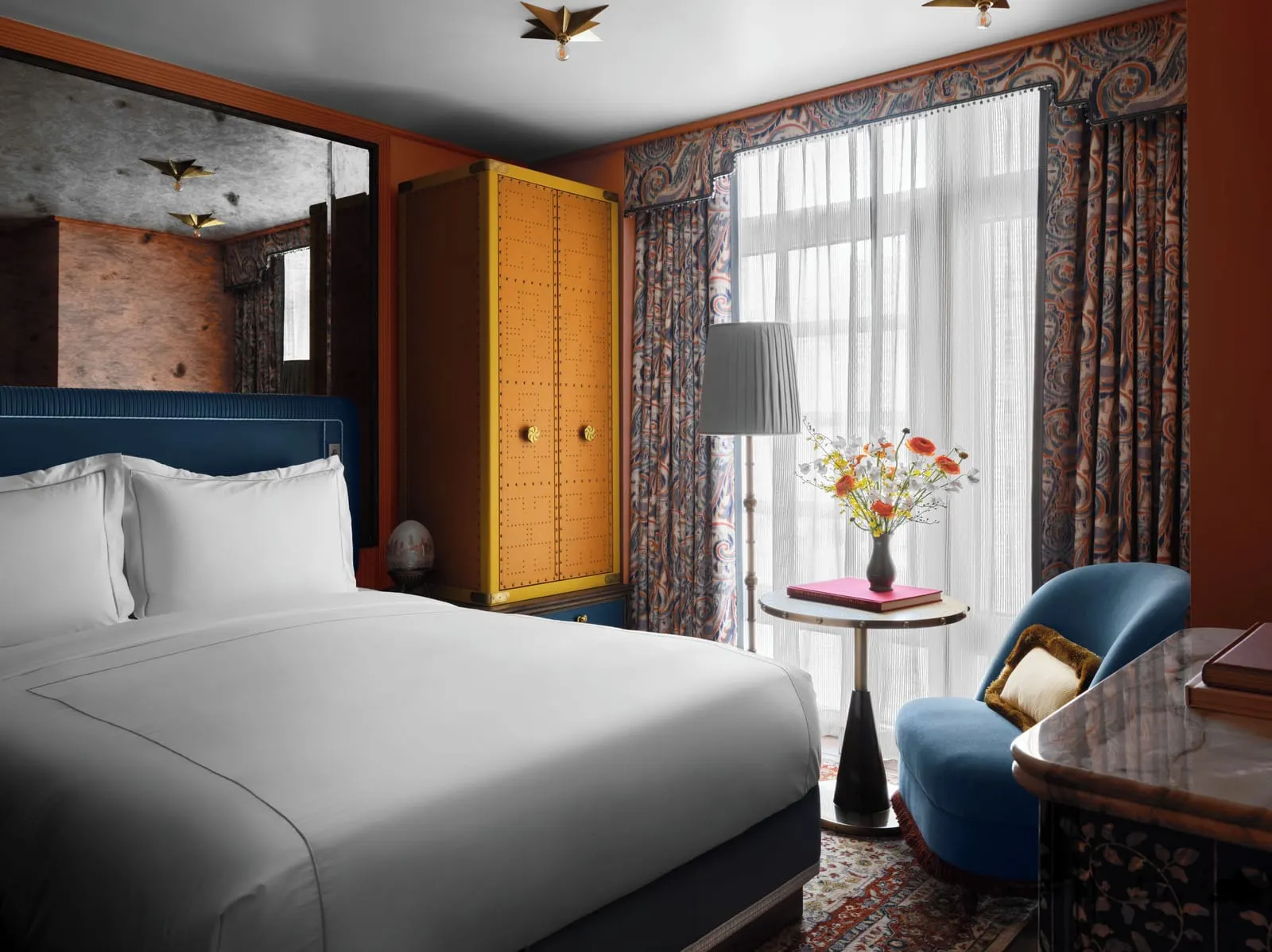 The NoMad Queen at The Fifth Avenue Hotel: a bed with white linen and a dark blue headboard beside a tall studded orange wardrobe, with a blue armchair and a round side table in front of a sheer curtained window