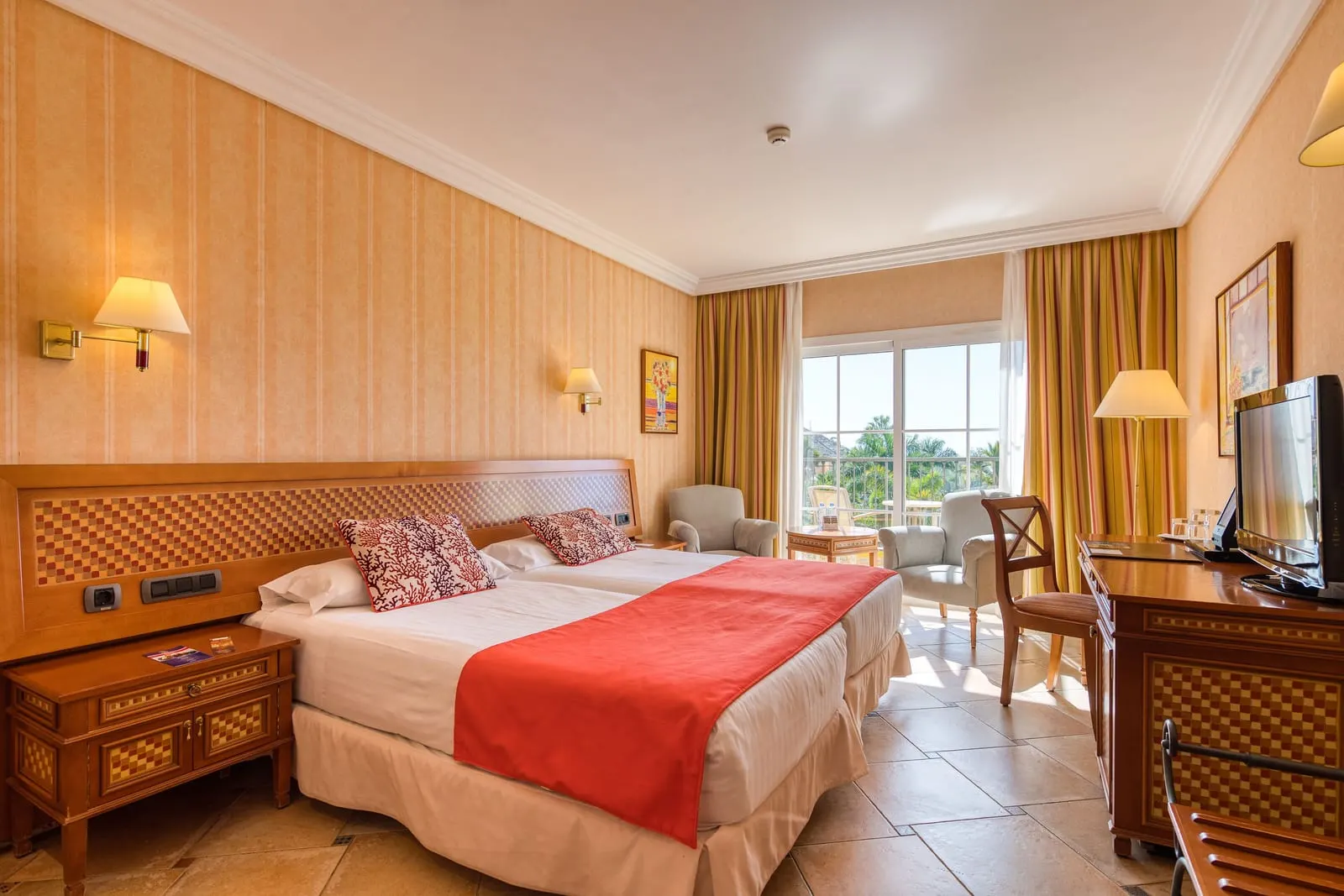 Double Room Pool View at Hotel Cordial Mogán Playa: two single beds side by side under wall lamps, a desk with a television, and two armchairs by glass doors to the balcony